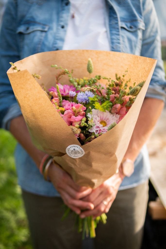 Mains d'une femme tenant un bouquet de fleurs dans les tons roses, dans un papier kraft