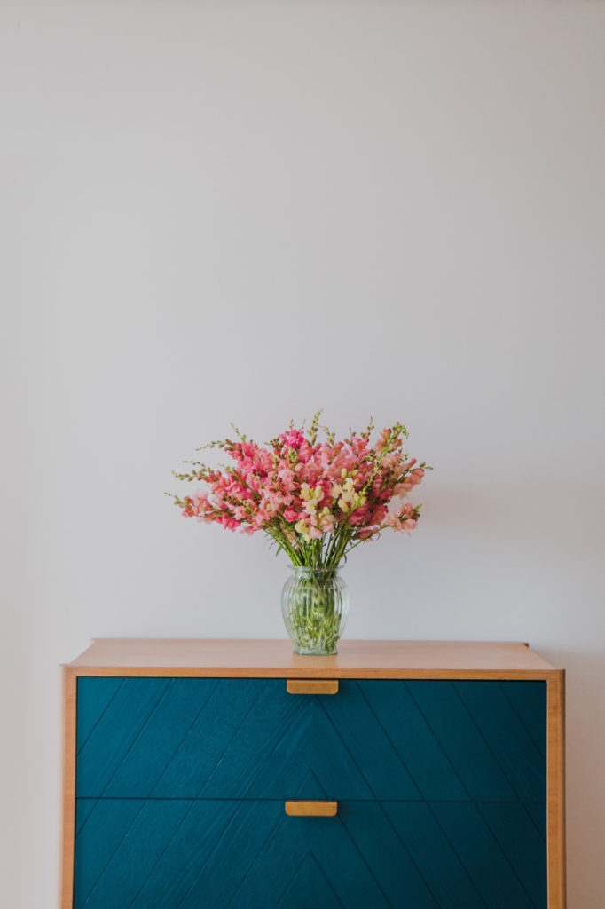 Bouquet de fleurs roses dans un vase transparent, posé sur un meuble de bois aux portes bleues